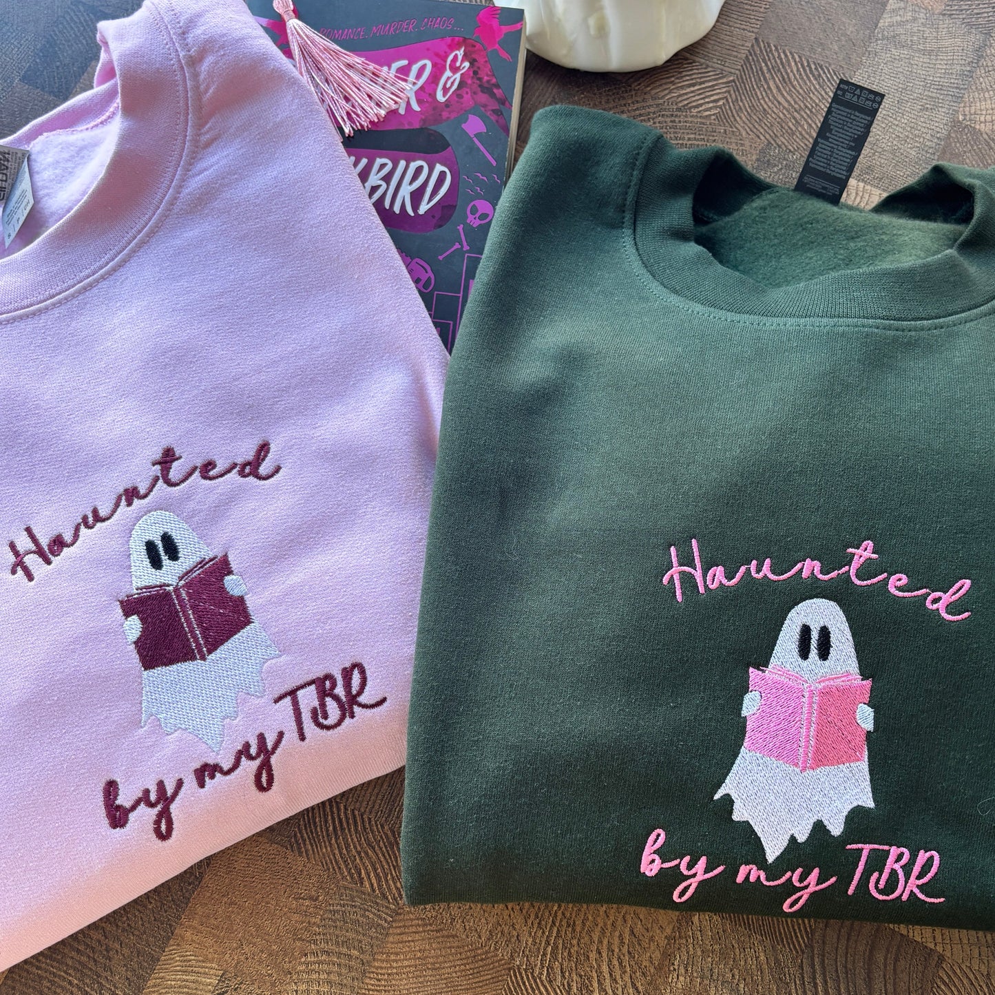 Two folded sweatshirts with a ghost reading a book design and 'Haunted by my TBR' text on a wooden surface.