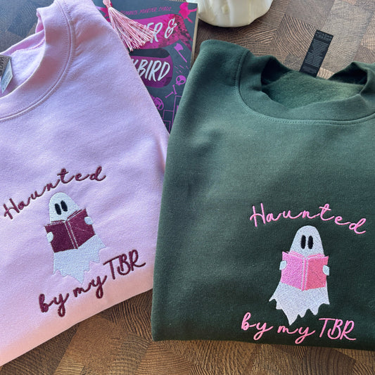 Two folded sweatshirts with a ghost reading a book design and 'Haunted by my TBR' text on a wooden surface.